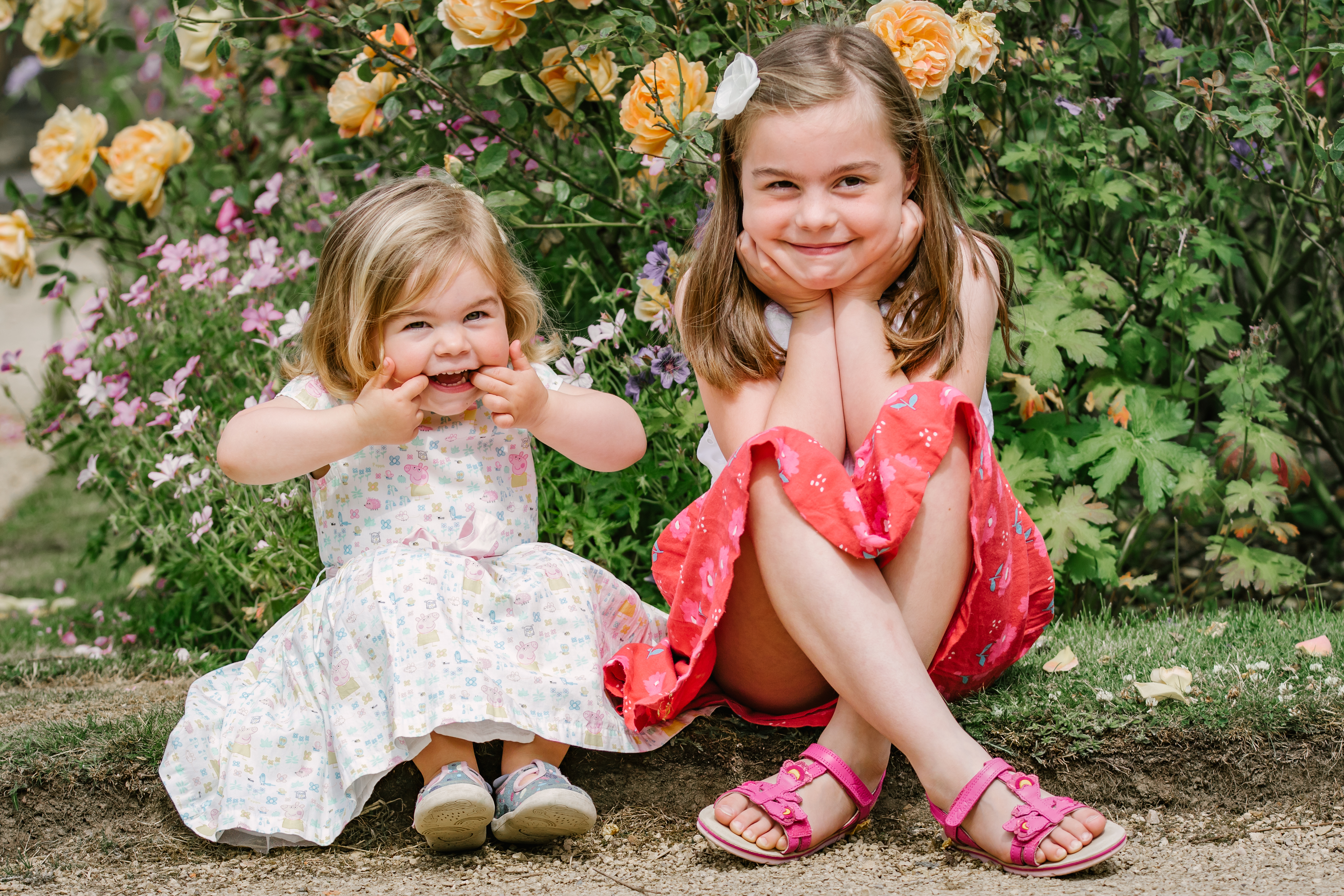 Two young sisters sitting together and pulling playful faces in front of a rose bush